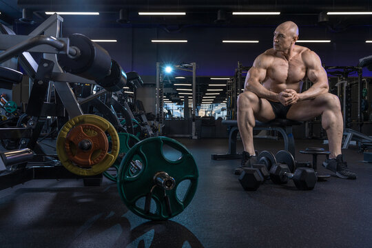 A muscular man sits on a weight bench in a gym, surrounded by equipment and dumbbells.