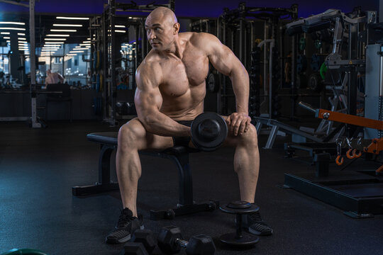 Muscular man rests on a weight bench in a gym, surrounded by equipment and showcasing his physique.