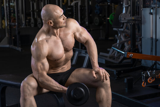 Muscular man rests on a bench in a gym, holding a dumbbell and gazing thoughtfully into the distance.