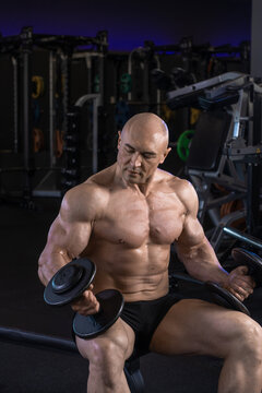 Muscular man lifts a dumbbell while seated in a gym with weightlifting equipment.