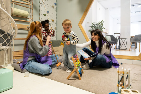 Teachers and toddlers are playing with toys and reading a book in a modern kindergarten classroom