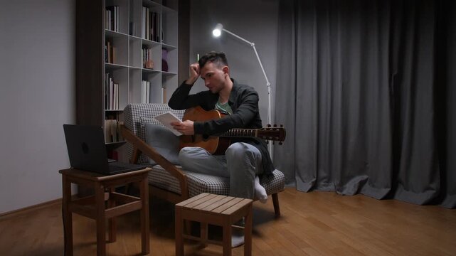 Man sits in cozy home studio at night, holding acoustic guitar and sheet music while laptop glows, focused on songwriting and creative practice with calm concentration