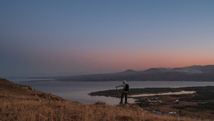 A hiker stands atop a hill overlooking Lake Sevan under a vast twilight sky. The stillness of the lake and distant mountain ranges creates a sense of quiet awe and connection with nature. © banphote