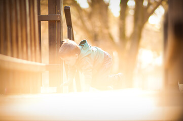 A child in a green jacket crawls excitedly on the ground, surrounded by the golden glow of the afternoon sun, exploring the wooden structure of a playground.