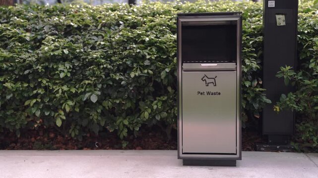 Modern stainless steel pet waste bin located in a public park next to green bushes. Features a dog icon and text for disposal of animal excrement to maintain cleanliness in outdoor spaces.