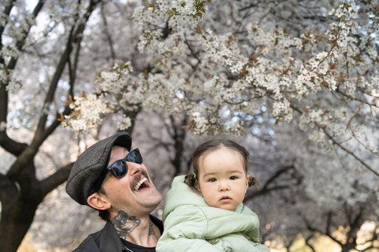 A young hipster father smiling portrait with his young daughter by the blossoming tree in the park on a spring day