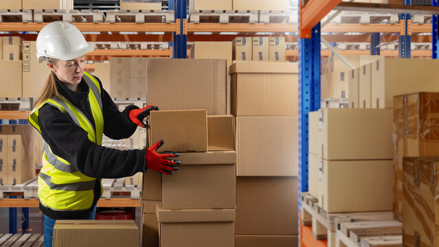 Warehouse operations, order fulfillment. Worker in helmet and safety vest stacks cardboard boxes on pallets, performing picking, packing, inventory handling, or distribution tasks.