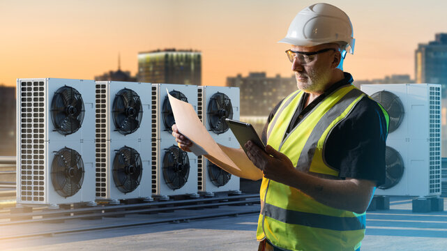 HVAC maintenance, Building services. Technician in hard hat inspects rooftop air conditioning units, comparing documents with tablet data to verify installation and system performance.