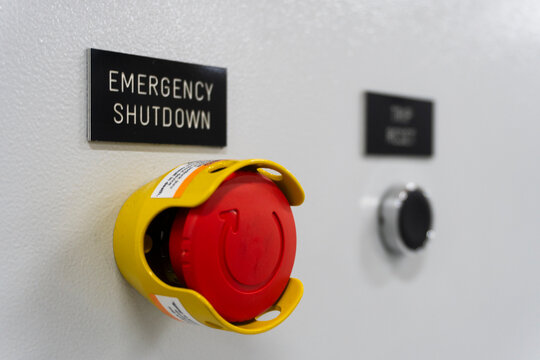 Red emergency stop button with yellow safety housing. Sign reads 'EMERGENCY SHUTDOWN'. Panel includes black push-button. Used for industrial safety or machinery control
