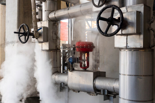 Steam escapes from industrial pipes and valves in factory setting. Metal pipes, red safety valves, and black handwheels dominate scene. Steam billows, suggesting active heating or processing systems