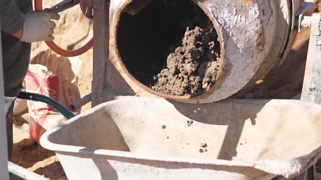 Heavy Concrete Flowing From Mixer Into Wheelbarrow, Operator Tilts Drum As Thick Gray Mix Drops And Fills Container, Shovel On Standby, Closeup Of Dense Texture During Pour.