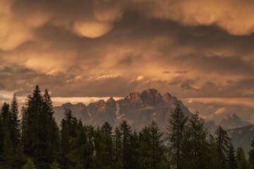 Obraz premium Majestic mountain peaks silhouetted against a dramatic orange sky filled with mammatus clouds and a dark pine forest foreground during a storm sunset.