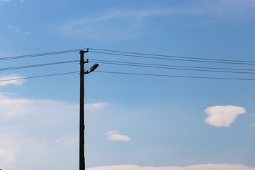Fototapeta premium Minimalist view of a silhouetted electric pole with electric wires and a street lamp against a bright blue sky with white clouds.