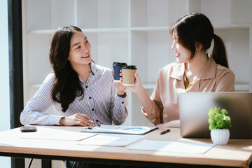 Fototapeta premium Two businesswomen reviewing charts and discussing ideas at a desk with a laptop and coffee, highlighting teamwork, data analysis, and modern office collaboration.