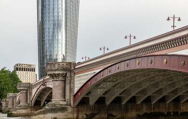 The ornate, scarlet-hued arches of Blackfriars Bridge sweep gracefully across the frame, their Victorian grandeur punctuated by golden medallions and classical stone piers. In the background, the shim