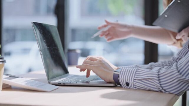 Close-up of two businesswomen working on a laptop, one pointing at the screen while the other types, showing teamwork, guidance, and collaboration in a bright office.
