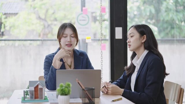 Two businesswomen discussing a house model and documents at a desk near a window, representing real estate consultation, property investment, and professional teamwork.