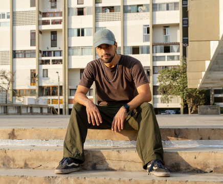 Urban Latino man sitting on concrete steps outdoors in casual streetwear in front of a residential building.