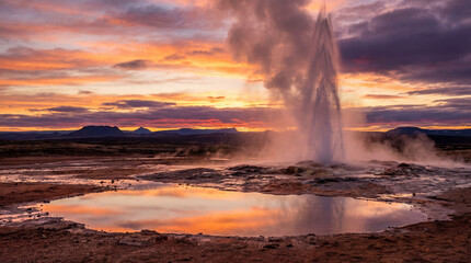 Fototapeta premium Geyser erupting with steam and water against a colorful sunset sky, reflecting in a nearby pool, surrounded by rugged terrain and distant mountains in Iceland