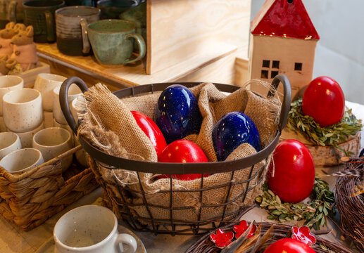 Metal basket lined with coarse burlap and filled with large glossy Easter eggs in deep red and dark navy blue. Cozy holiday still life with rustic ceramics, nest wreaths and decorative accents for spr