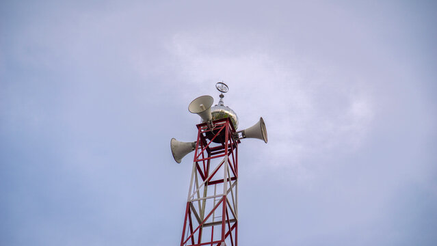 Red and white metal tower structure featuring loudspeakers and a small Islamic dome against a cloudy sky background - Menara pengeras suara masjid