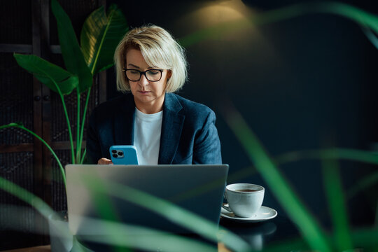 Stylish 50's confident mature businesswoman, middle aged company ceo director, experienced senior female professional, business coach using phone and working on laptop in office. Female leader