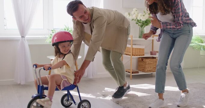 Family at home as daughter rides tricycle. Smiling, happy parents guide the child in a living room, the girl wearing a helmet for safety and balance. Bonding, leisure, fun and supportive play.