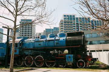 Old blue steam locomotive displayed near modern residential buildings in Belgrade. Concept of history, industry and urban development contrast © Ekaterina