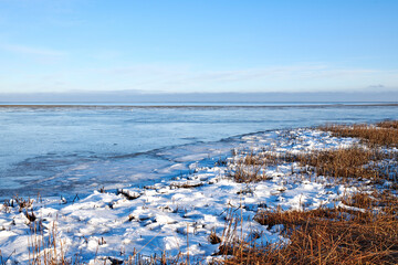 Naklejka premium Landscape, frozen and lake with grass in nature for climate change, winter season and morning frost. Space, blue sky or river with ice formation, snowfall field and icy conditions from global warning