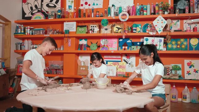Diverse high school people working on dough while asian girl modeling clay at pottery workshop. Group of happy diverse children working or making cup of clay. Creative activity concept. Edification