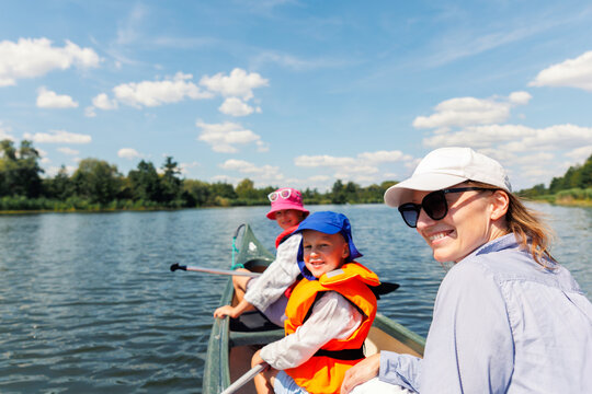 Woman two children enjoy having fun paddling canoe rent across lake river water wearing bright orange life vests hot sunny summer day. Healthy recreational activity lifestyle vacation trip journey