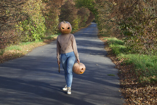 Creepy pumpkin head character walking on autumn road with axe and jack-o-lantern, cinematic horror concept for halloween, mysterious slasher figure in fall forest landscape