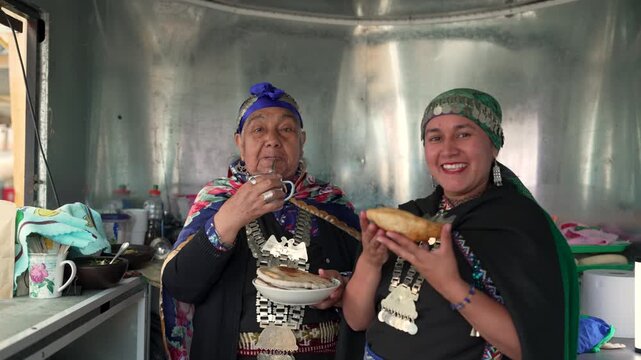 Two indigenous mapuche women in traditional clothing sharing sopaipillas and mate in a food stall