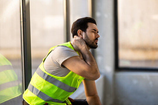 Man wearing a safety vest sits and holds his neck at a work site during the day while taking a break