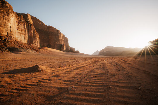 Early morning light bathes a red sand desert track in warm golden tones, with long shadows cast across towering sandstone cliffs shortly after sunrise, Wadi Rum, Jordan.