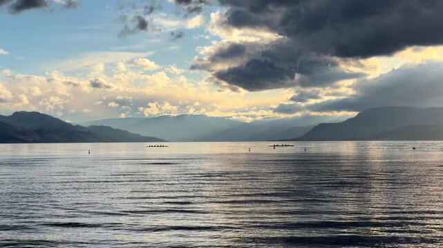Two dragon boats in the distance paddle across scenic lake with mountains on a cloudy day