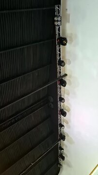 A row of moving head LED spotlights flash in sync on a metal truss mounted in a venue. The lighting setup is designed for a performance with dynamic effects.
