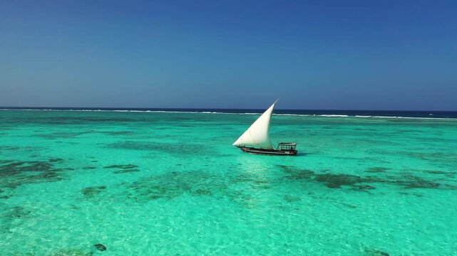 A sailboat moves through blue waters with a white sail raised. Other boats are visible in the distance. The sky has some clouds and the sun is shining bright.
