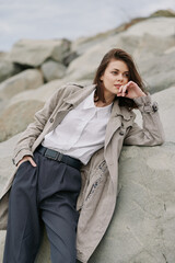 Young woman in textured coat and white shirt leans on rock outdoors, looking sideways with...