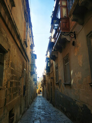 Obraz premium Narrow Maltese alley between weathered stone buildings under clear daylight. The image conveys the concept of historic urban space and Mediterranean street scenery.