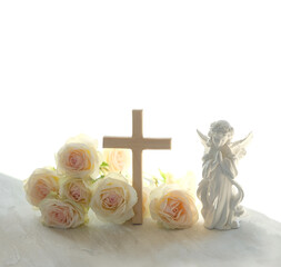 Naklejka premium Easter holiday background. Beautiful angel figurine with wooden cross and rose flowers on light table close up. symbol of Easter, Christianity religion, faith in God, prayer, memory. soft focus