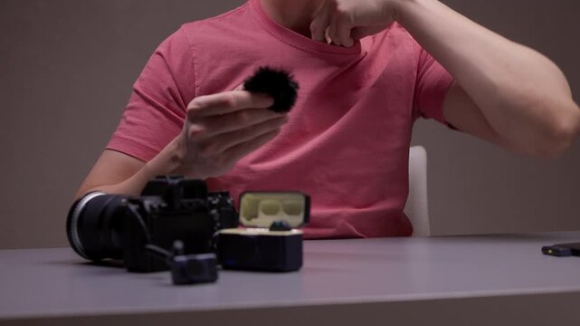 Professional content creator sitting at desk and attaching wireless lavalier microphone with furry windscreen to t-shirt, preparing equipment for video recording session in dark studio, close-up.