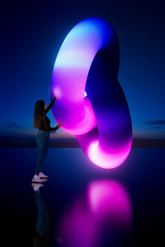Latin woman touching glowing 3D abstract loop structure with gradient light in dark environment