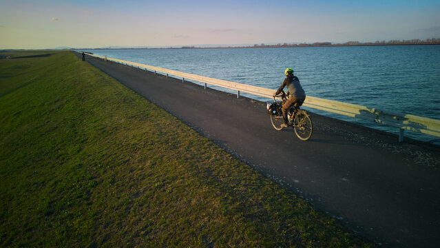 Aerial view of bicycle tourist with panniers. Bikepacking on a long, straight Danube bike path near Bratislava, Slovakia at sunset. Lone rider on dike trail.