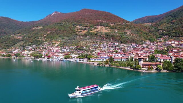 Italy Lake Iseo Predore village aerial drone tracking shot of boat departing from pier. Medieval castle tower and scenic mountains landscape. Bergamo province travel scenery 4k video Lombardy