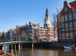 Obraz premium Classic Amsterdam Canal and Montelbaanstoren - Iconic Amsterdam canal scene featuring a bridge, historic gabled houses, and the distinctive Montelbaanstoren tower under a clear blue sky.