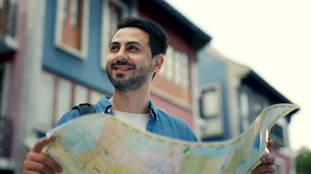 Portrait of happy brunet man plans trip reading map in town district. Bearded male tourist in casual attire chooses place to visit on excursion in old city