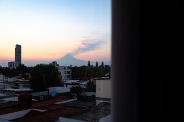 Silhouette of Popocatepetl volcano emitting smoke against vibrant sunset sky seen through dark curtain from interior space in Puebla, Mexico © simonmayer