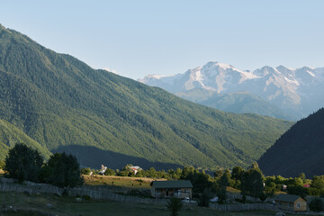 Fototapeta premium A tranquil mountain valley scene unfolds with green hills, distant snow capped peaks, and a clear blue sky. Meadows dot the landscape as rustic cottages sit beside stone walls and gentle sunlight