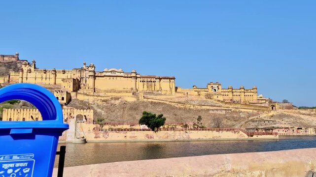 A bright daytime  showing the grand Amer Fort spread across the Aravalli hillside above Maota Lake, with yellow sandstone walls, palace structures.